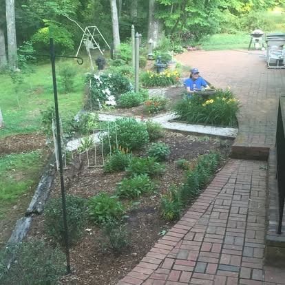 A person in a blue shirt sits on a stone birdbath in a lush garden alongside a brick patio and walkway.