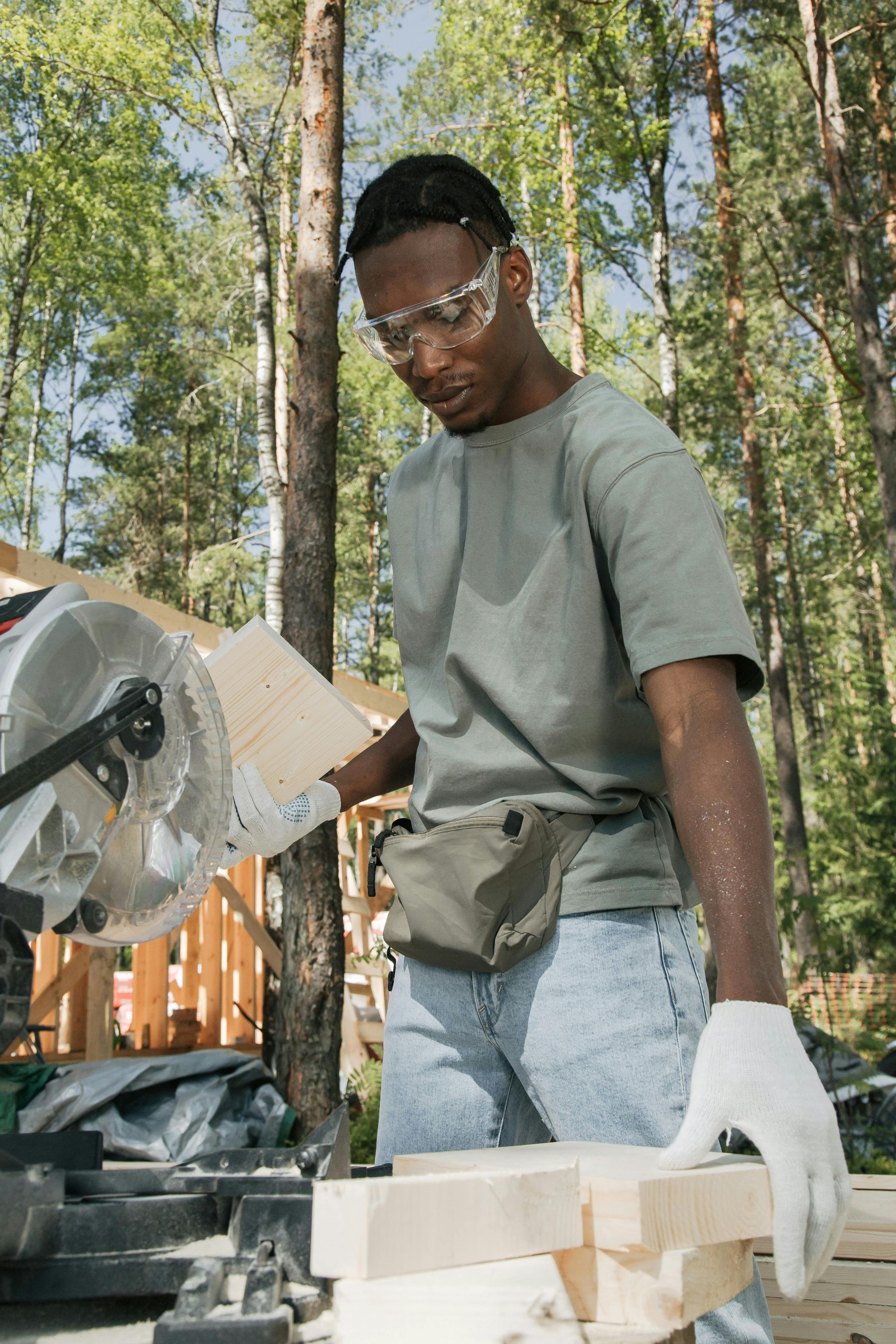 A person wearing protective goggles and gloves operates a miter saw to cut a wooden board outdoors in a forest.