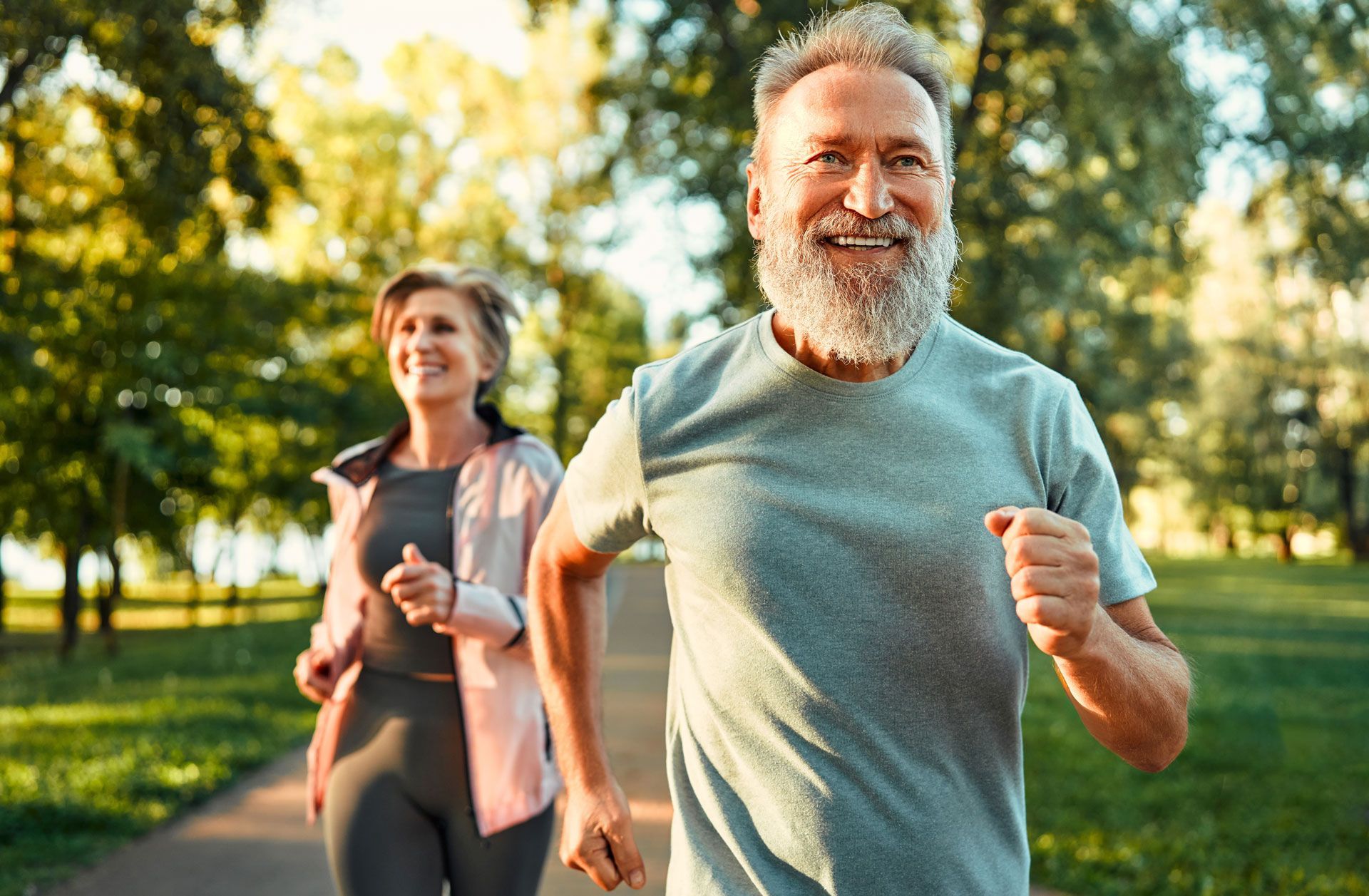 Two people running on a path in a park; man with grey beard smiles, woman follows behind.