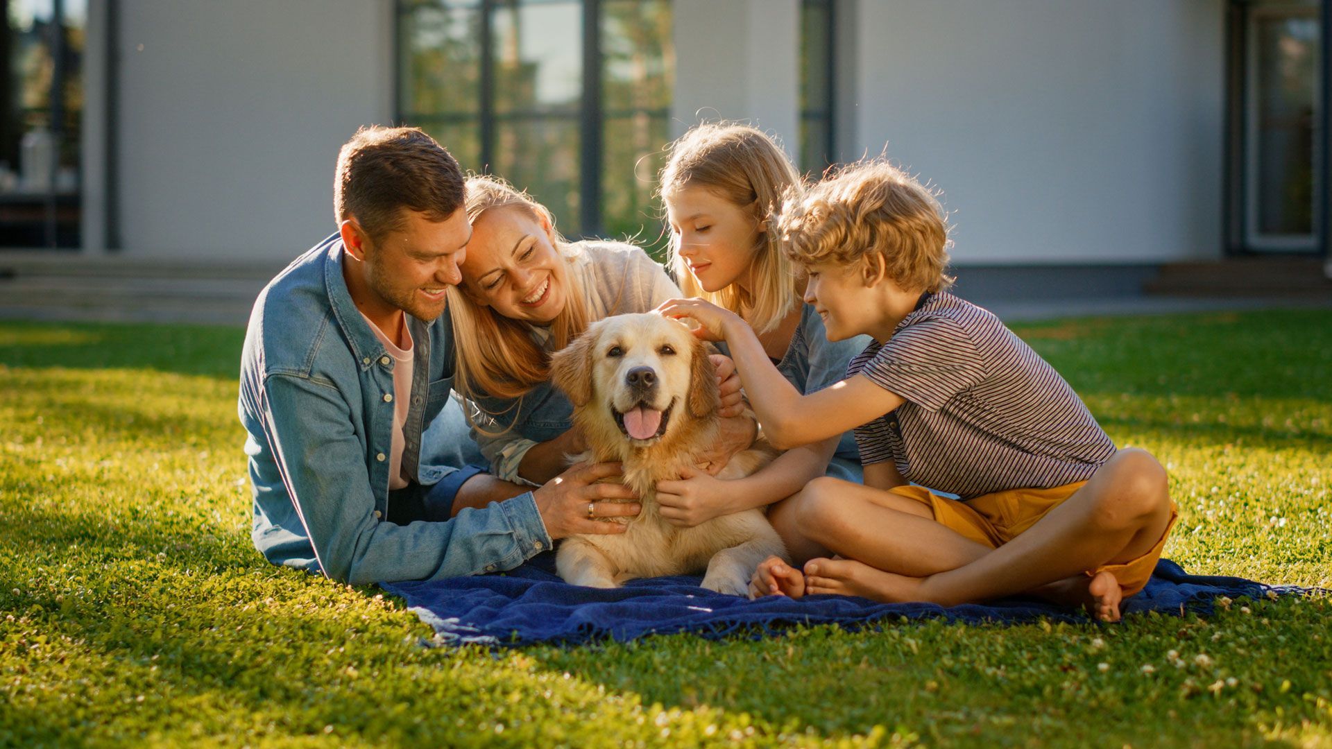 Family pets a golden retriever on a blanket in a sunny yard; they are petting the dog.