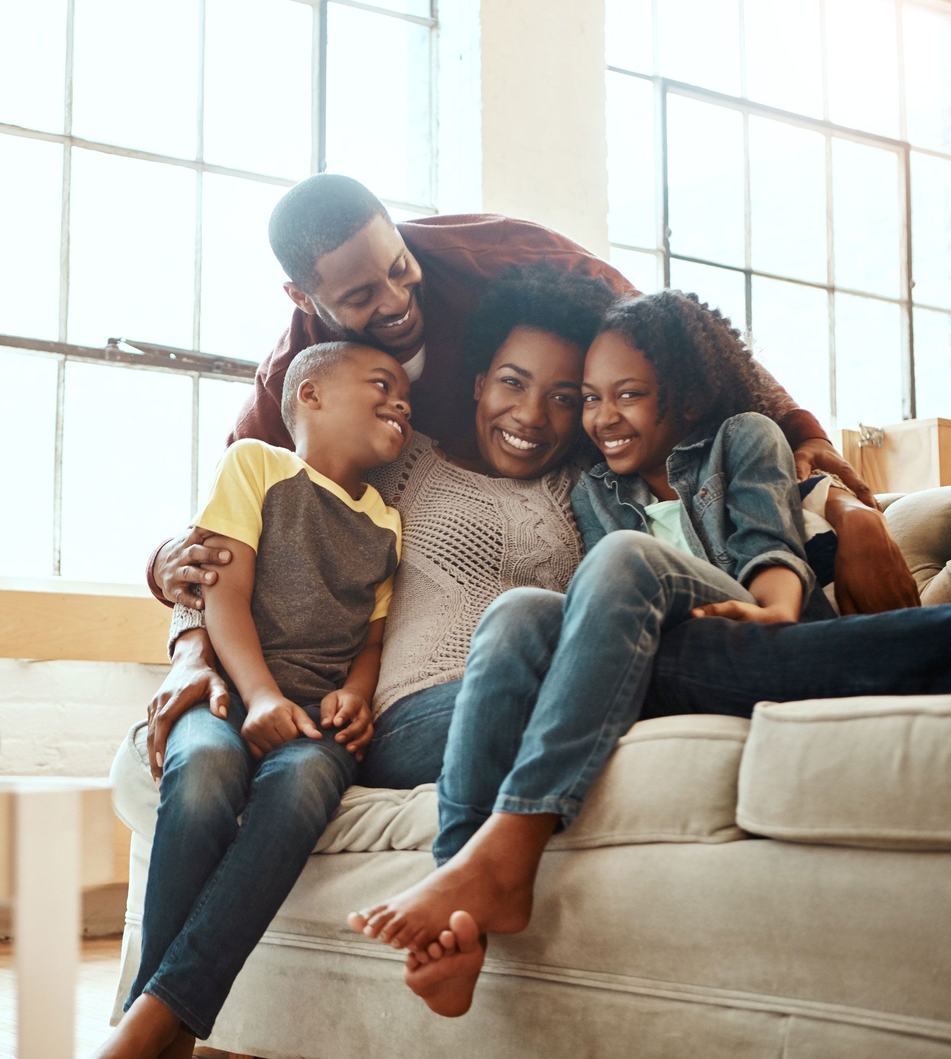 Family of four smiling and cuddling on a sofa near a large window.