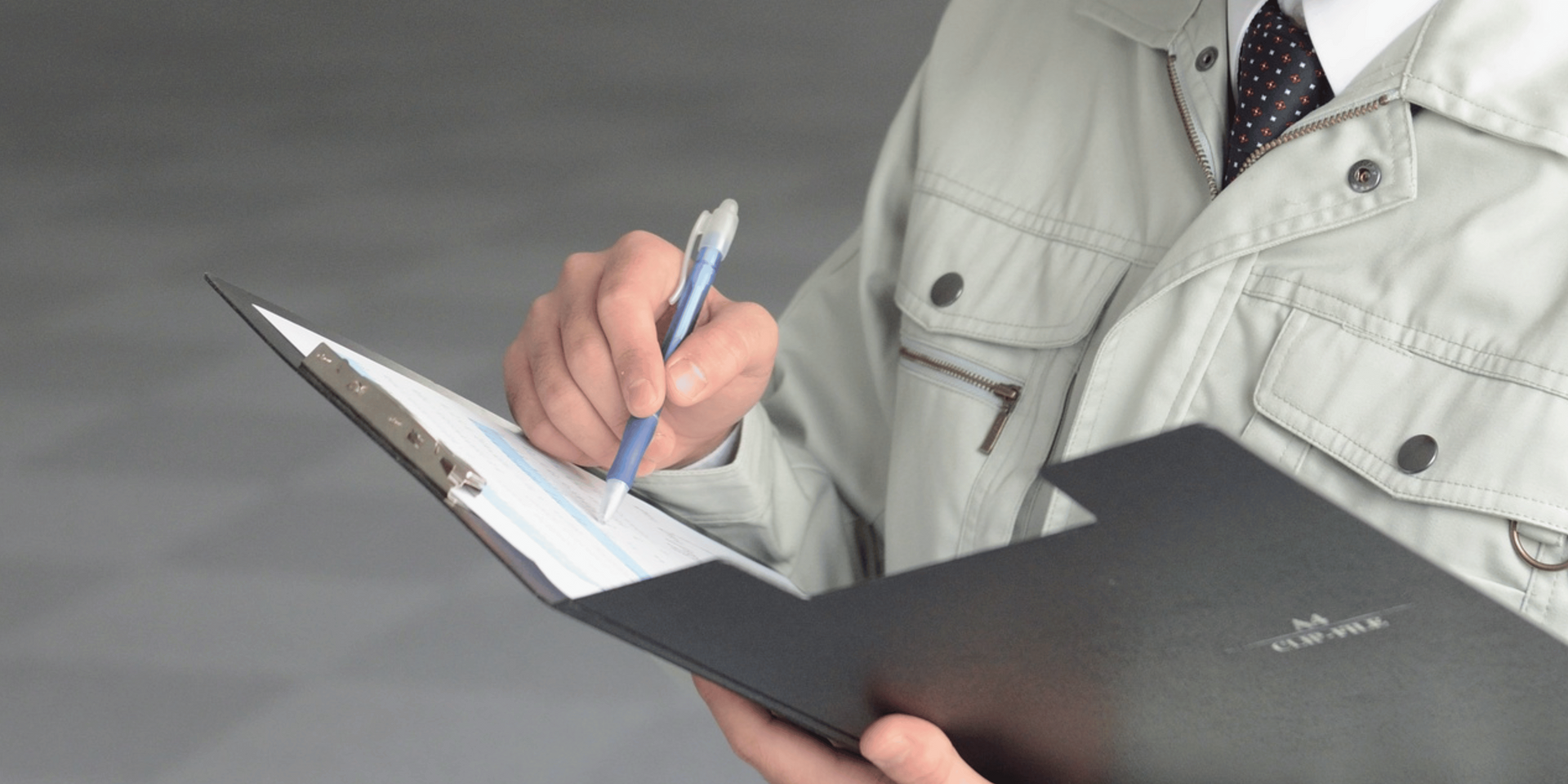 Person's hand writing on a clipboard with a blue pen. The background is a bathroom with a mirror and sink.