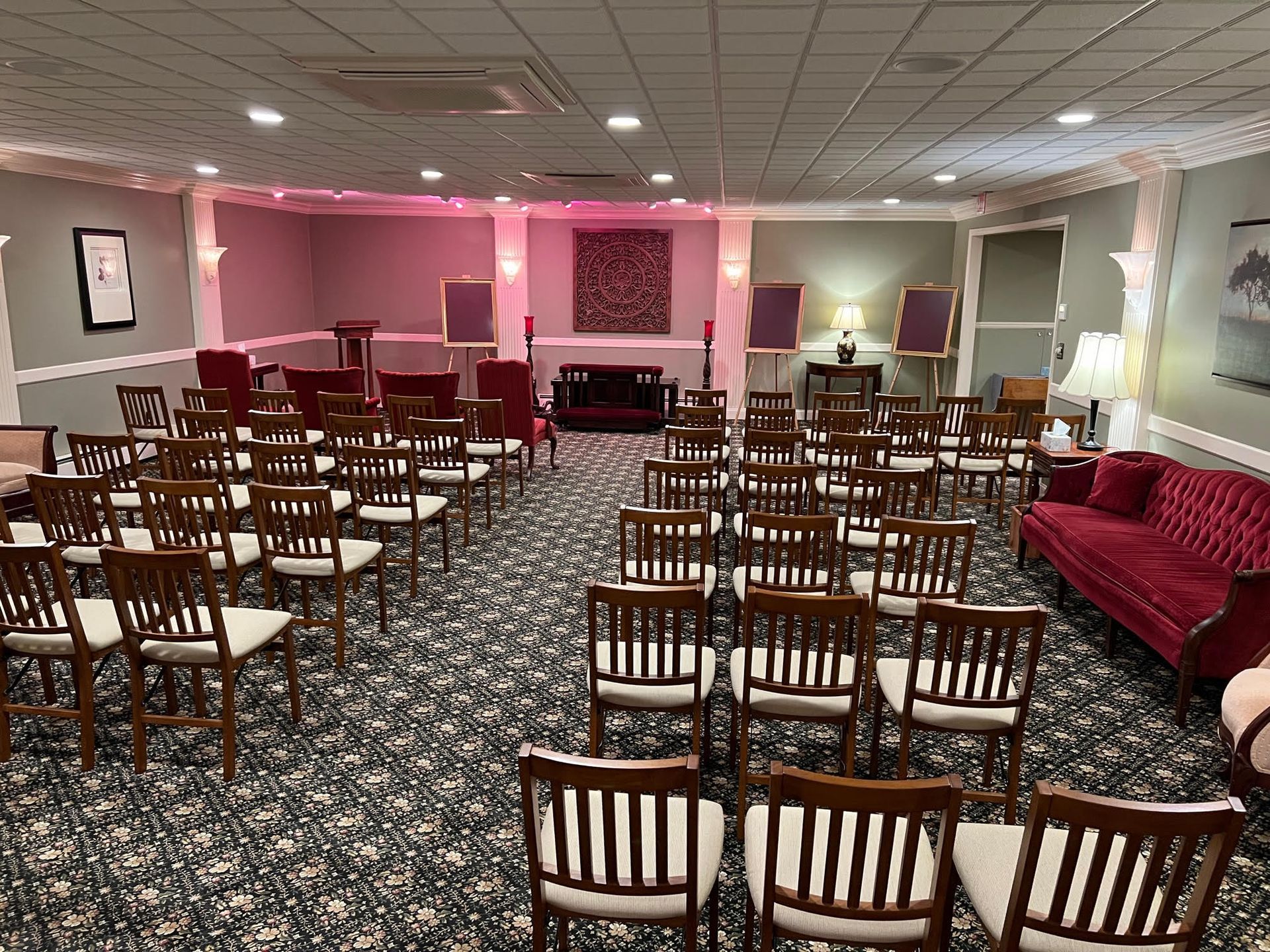 A carpeted funeral home viewing room with rows of wooden chairs facing a central podium and floral wall hanging.