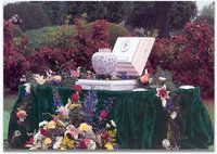 A funeral urn sits on a table draped in green cloth, surrounded by colorful flowers, set outdoors against red bushes.