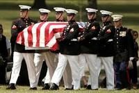 US Marines in dress uniforms carry a casket draped in an American flag during a military funeral service on grassy land.