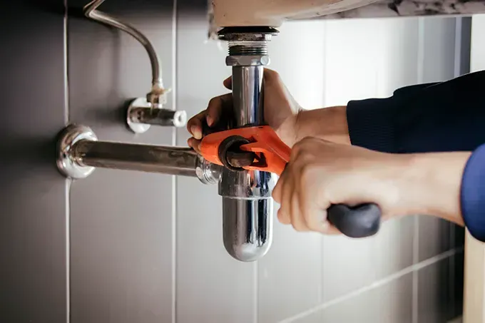 Hands using a wrench to tighten a chrome sink pipe under a bathroom basin