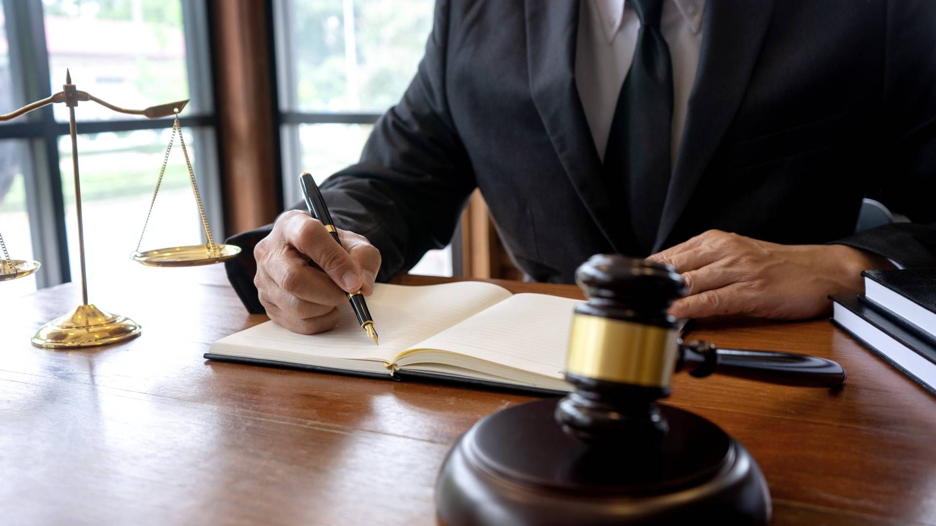 Lawyer writing in a notebook next to a gavel and scales of justice on a wooden desk.