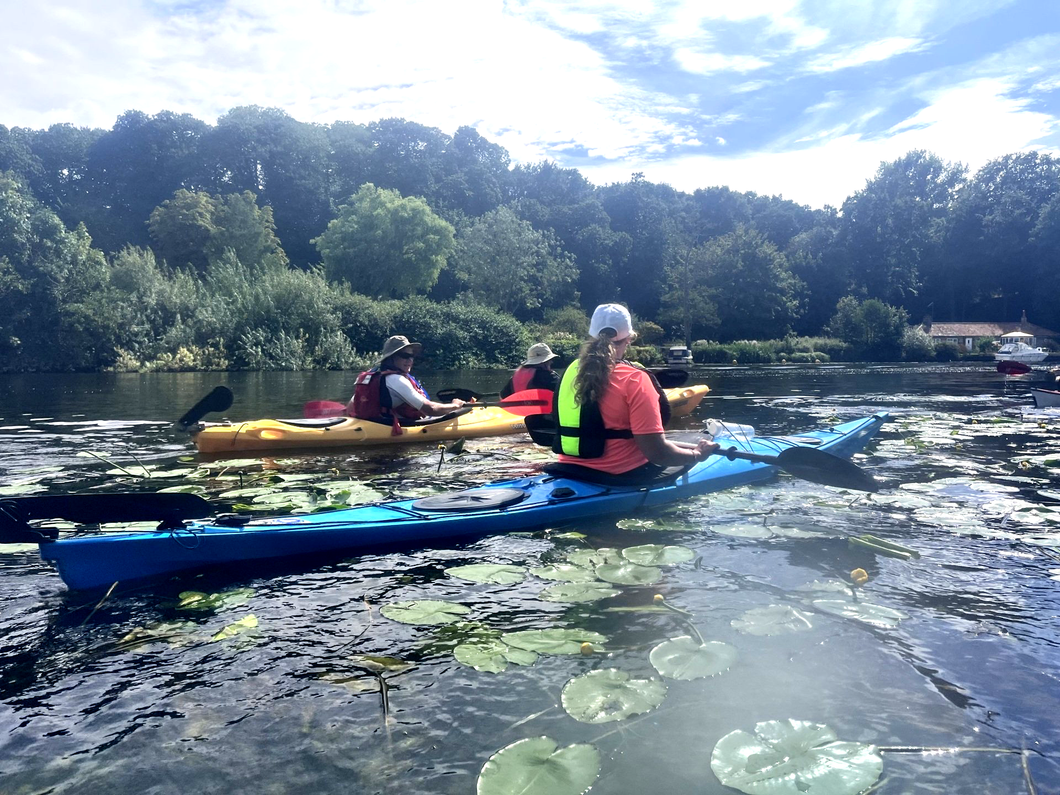 Kayakers paddling on a lake. Trees line the background. The sky is partly cloudy.