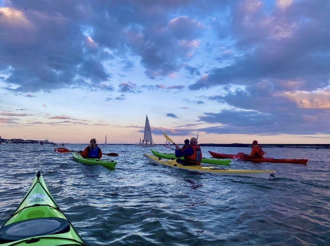 Kayakers on water at sunset, silhouetted against a cloudy sky. Sailboat in the distance.