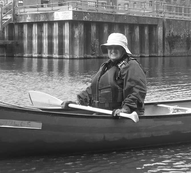 Woman wearing a hat and sunglasses, smiling, in a life vest, with two partially visible people.