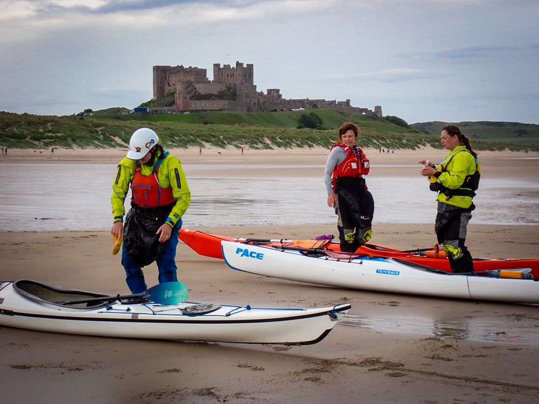 Three people preparing kayaks on a sandy beach, castle in background.