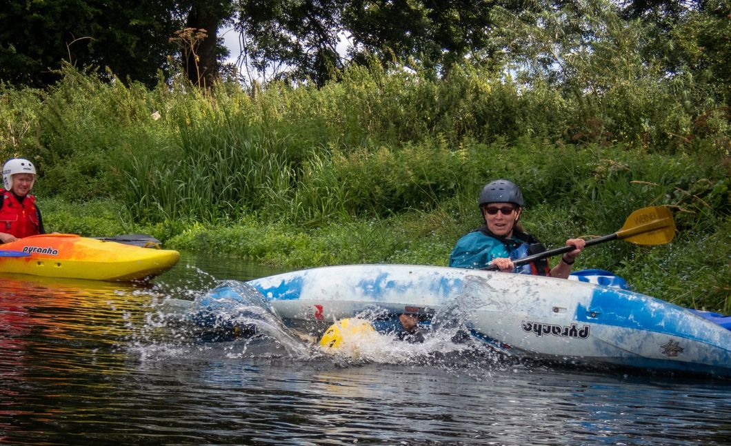 Two kayakers on a river. One in white and blue kayak is splashing water. Other kayaker in yellow kayak. Green foliage in background.