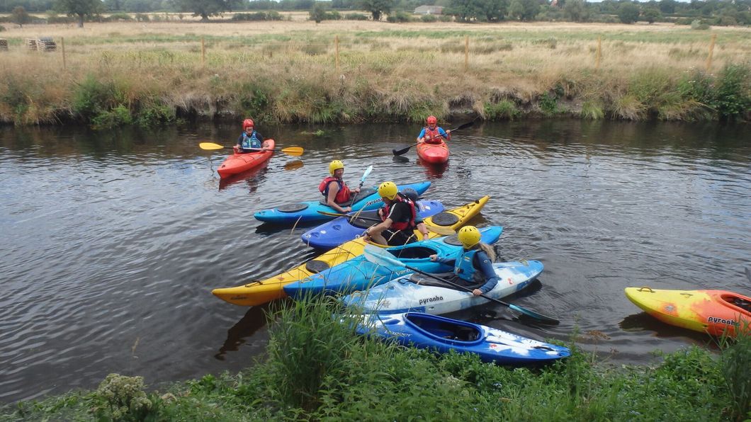 Kayakers in colorful boats on a calm river. They wear helmets, paddles in hand, near grassy banks.