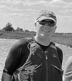 Man in life jacket smiles at camera, wearing a dark collar, outdoors.