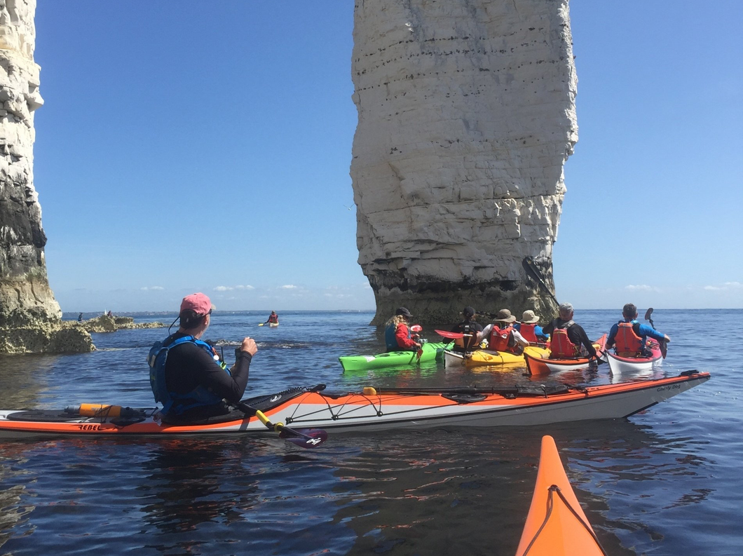 Kayakers near white cliffs under a blue sky. Some are in a multi-person kayak; other individual boats.