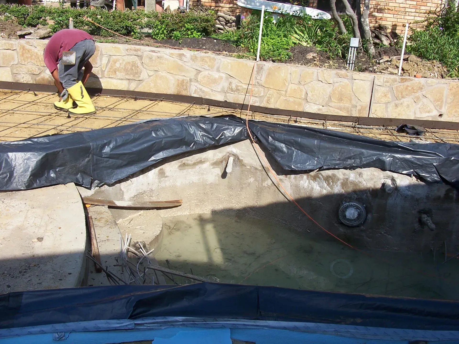 A person in yellow boots works on a drained pool, partially covered with black tarp. The setting is outdoors, with a stone wall in the background.