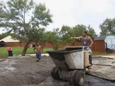 Two men pouring concrete with a motorized wheelbarrow in a residential yard. One points, the other watches.