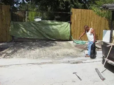 Man using a hose on a concrete surface in a yard, near a wooden fence and a tarp.