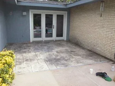 A concrete patio with patterned paving stones, adjacent to a house with blue siding and brick wall, leads to glass French doors.