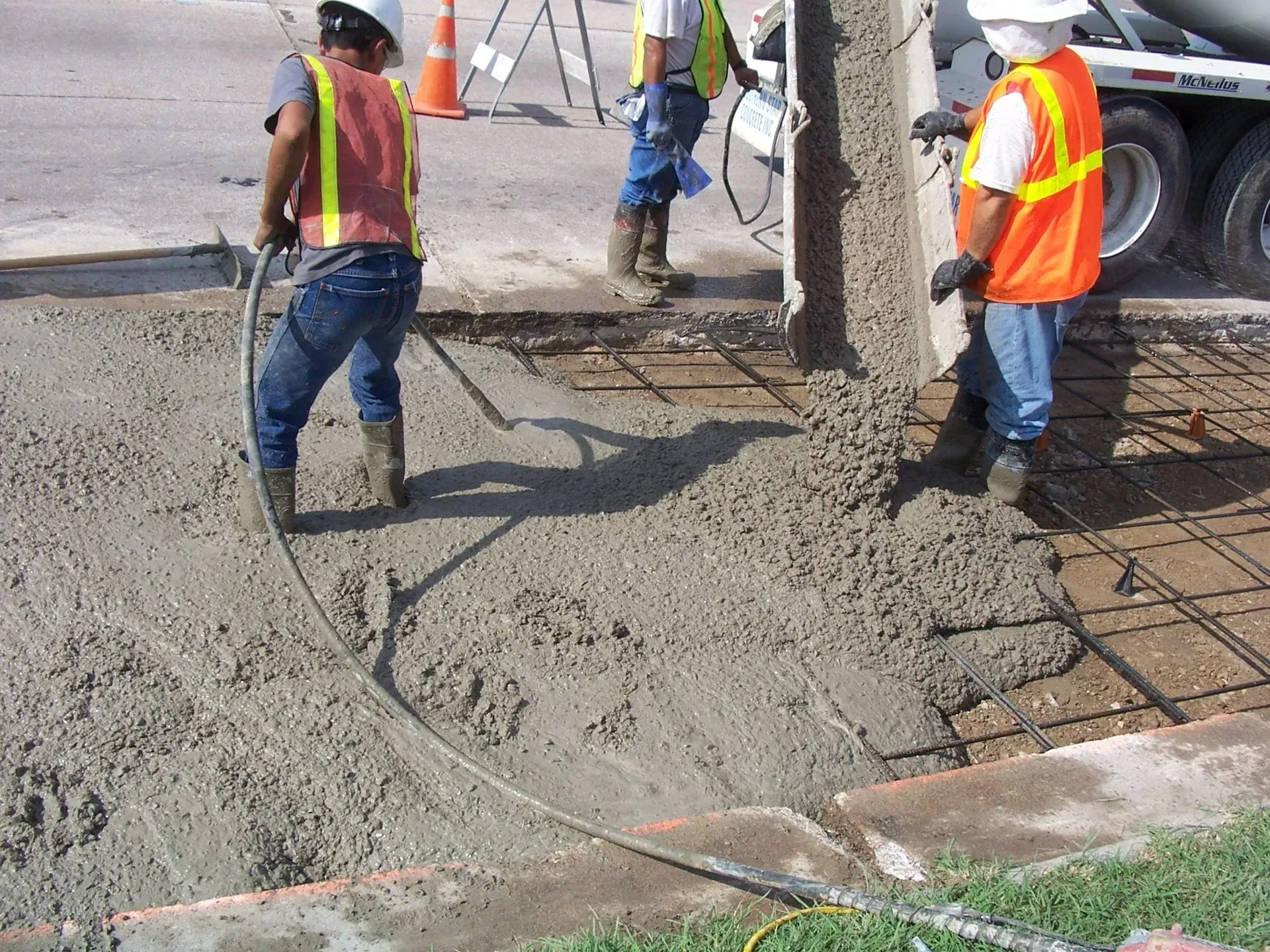 Construction workers pouring concrete onto a rebar grid at a sidewalk, using hoses connected to a truck.