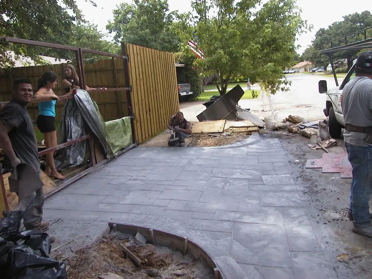 Construction workers laying concrete pavers on a driveway. Two women and a man are nearby, and a truck is parked in the background.