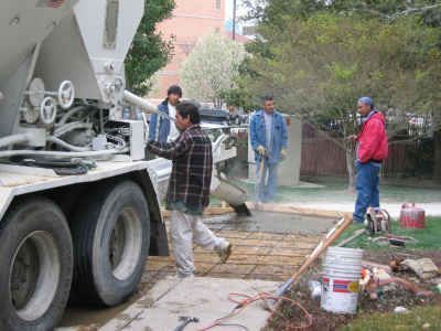 Construction workers pouring cement from a truck onto a rebar grid; a residential setting.