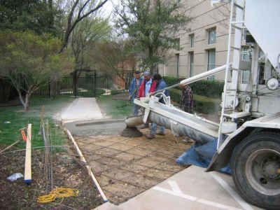 Concrete being poured from a truck onto a prepared area with rebar, sidewalk in progress. Workers are present, a building in the background.