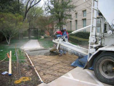 Concrete being poured for a walkway. Workers in red are guiding a cement truck's chute over rebar on the ground next to a finished sidewalk.
