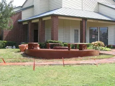 A brown concrete seating area with tables and pillars in front of a building with red brick and white siding.