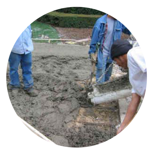 Construction workers pouring concrete on a project. The men are wearing blue work clothes. One worker holds a shovel, another is guiding the concrete, and a third is standing.