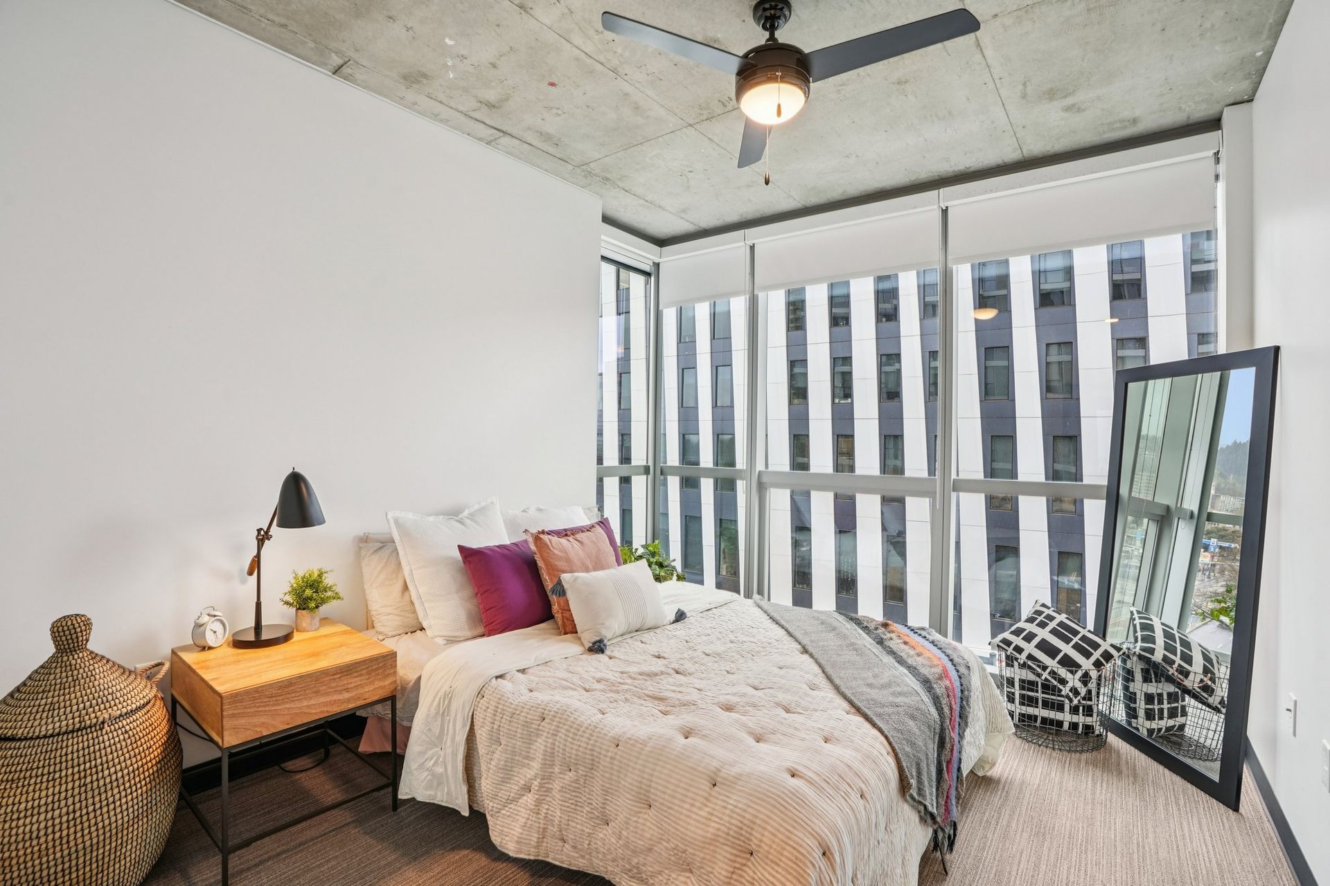 Modern bedroom with bed, nightstand, floor mirror, and large windows with city view