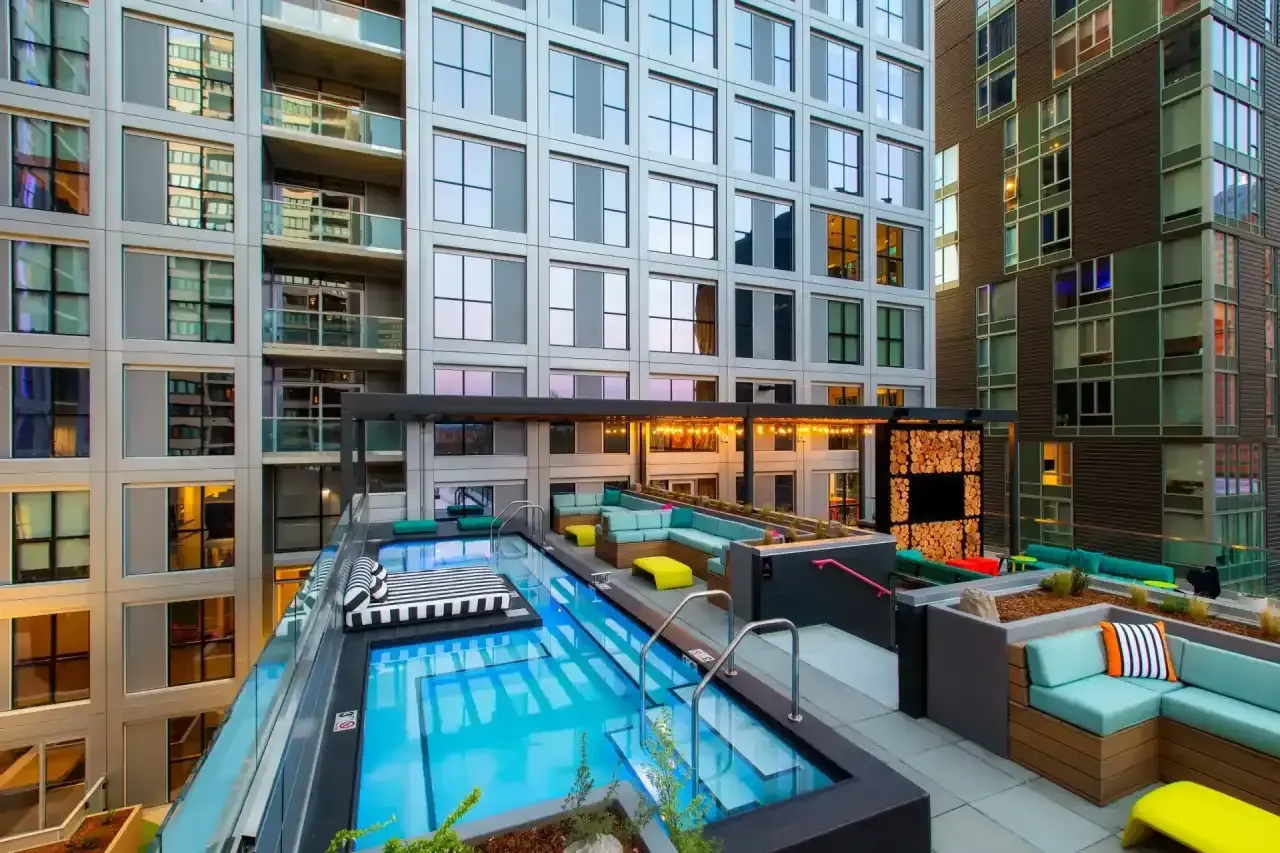 Rooftop pool deck with pool, seating, and high-rise buildings in the background. Turquoise, yellow, and black colors.