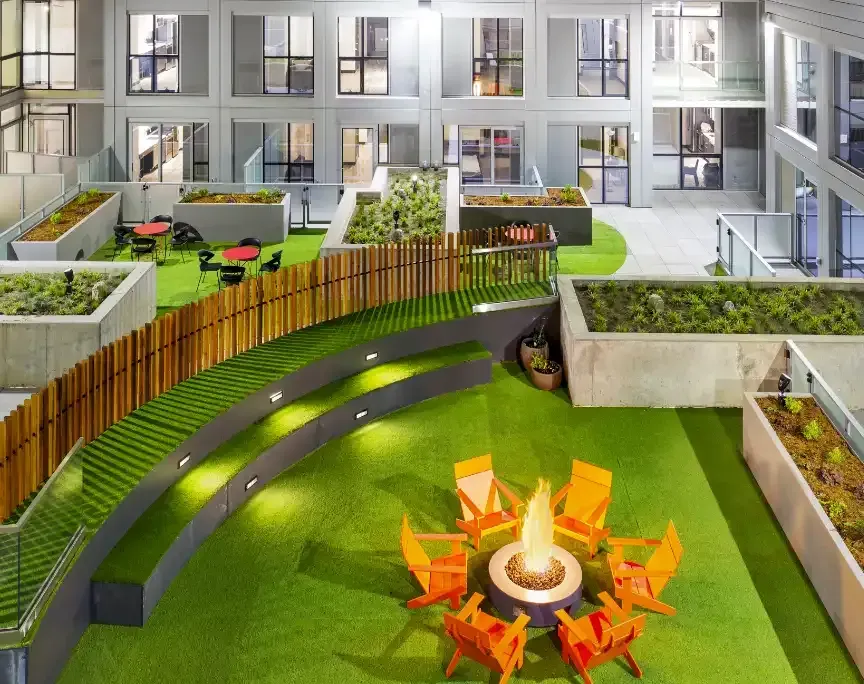 An outdoor rooftop lounge at night with a fire pit, seating, and garden areas surrounded by buildings.