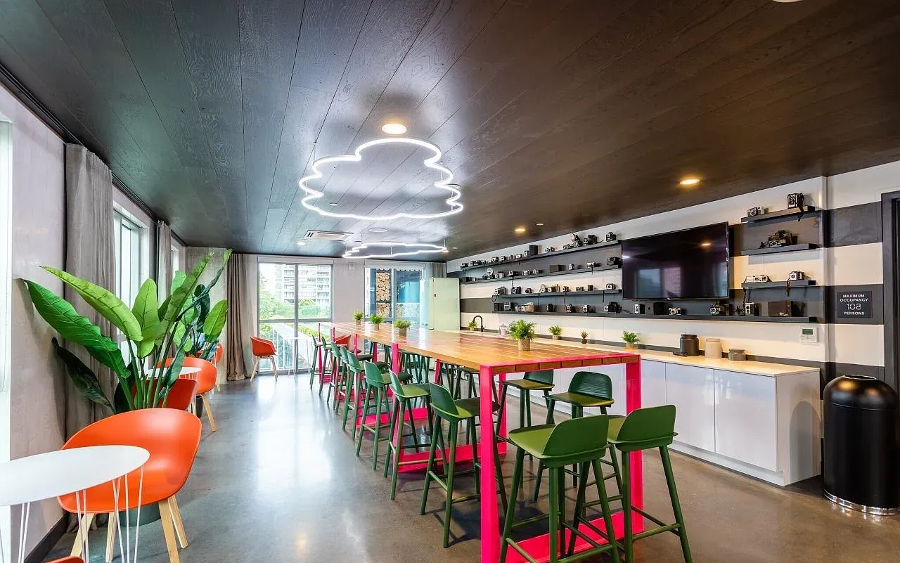 Long, narrow room with a pink table and green stools. Orange chairs with plants and a TV with shelves. Black ceiling.