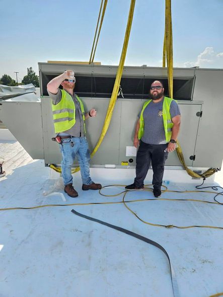 Two workers in safety vests standing on a snowy rooftop beside a large HVAC unit and yellow lifting straps.