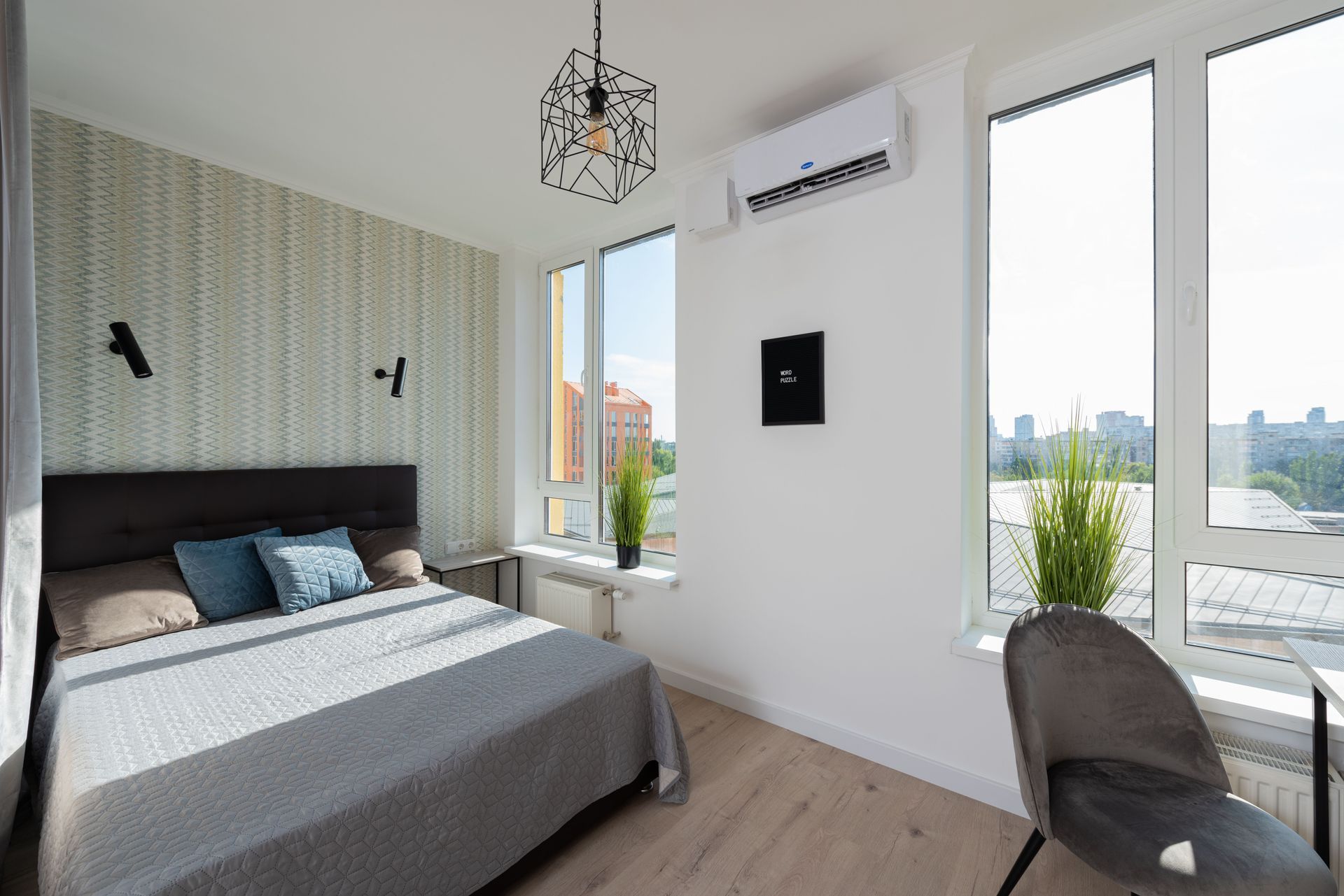 Modern bedroom with gray bed, patterned accent wall, white walls, large windows, and city view.