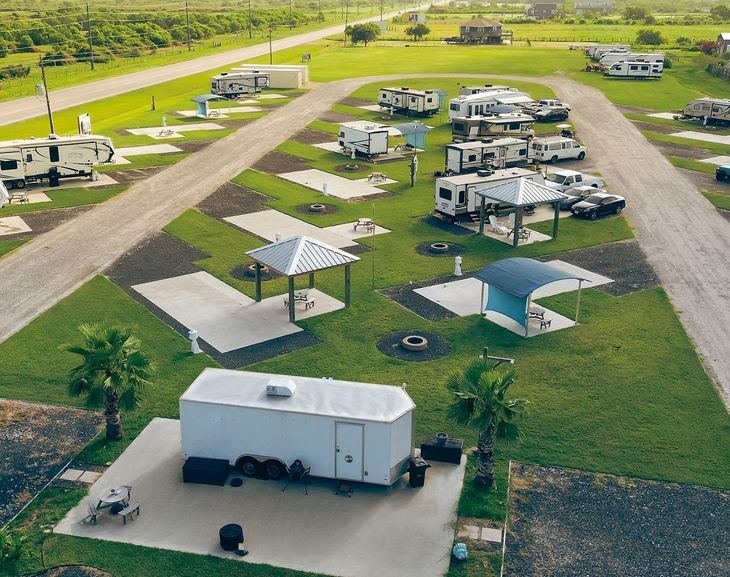 RV park with campers parked on grass lots. Shelters and palm trees dot the area.