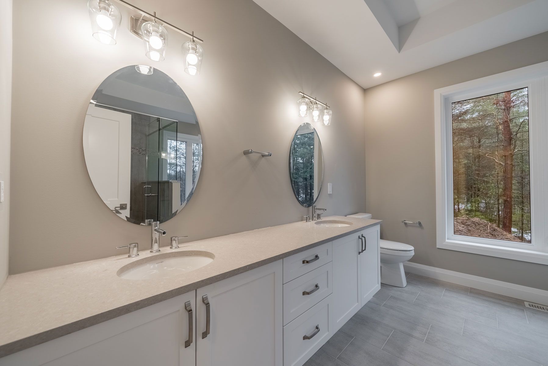 A bathroom with two sinks , two mirrors , a toilet and a window in Fidelity Homes Signature build in Cramahe Township