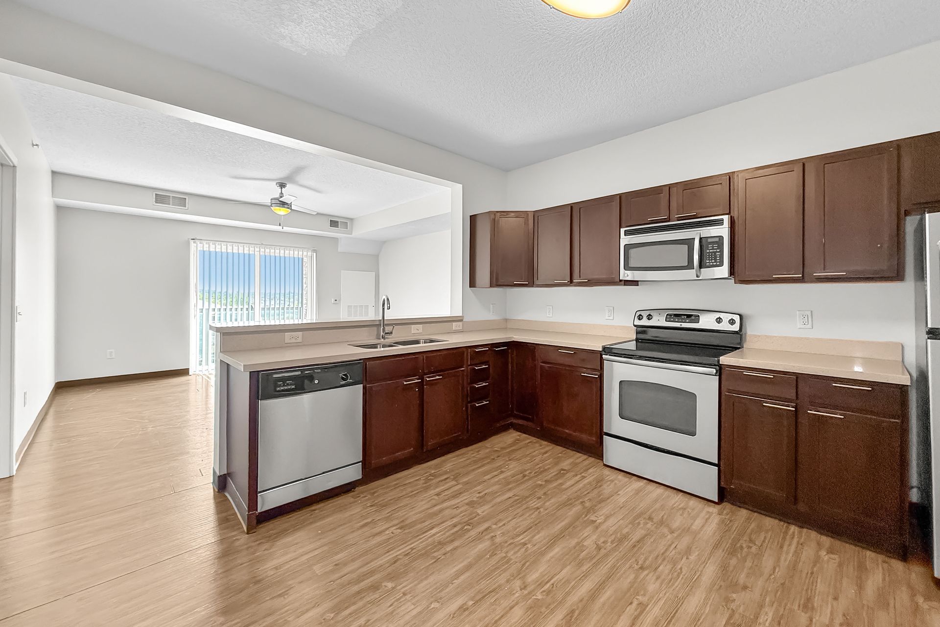 Living room with brown sofa, coffee table, and open kitchen with bar stools. Hardwood floors and white walls.