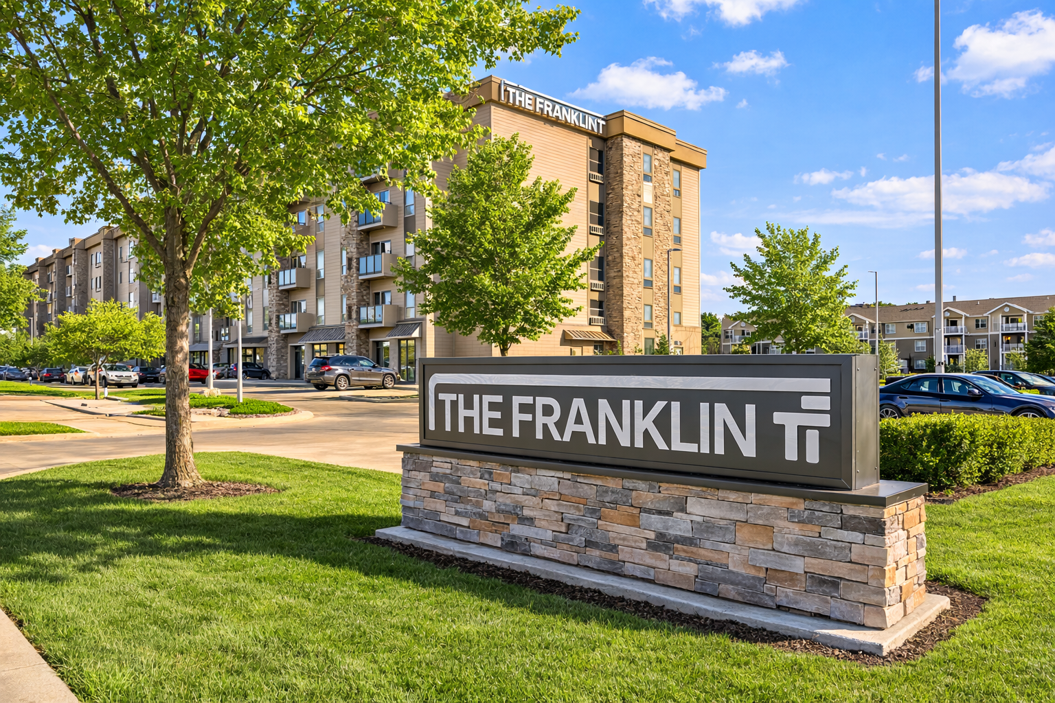 Apartment building with stone accents, balconies, and parked cars on a grassy lawn under a blue sky.