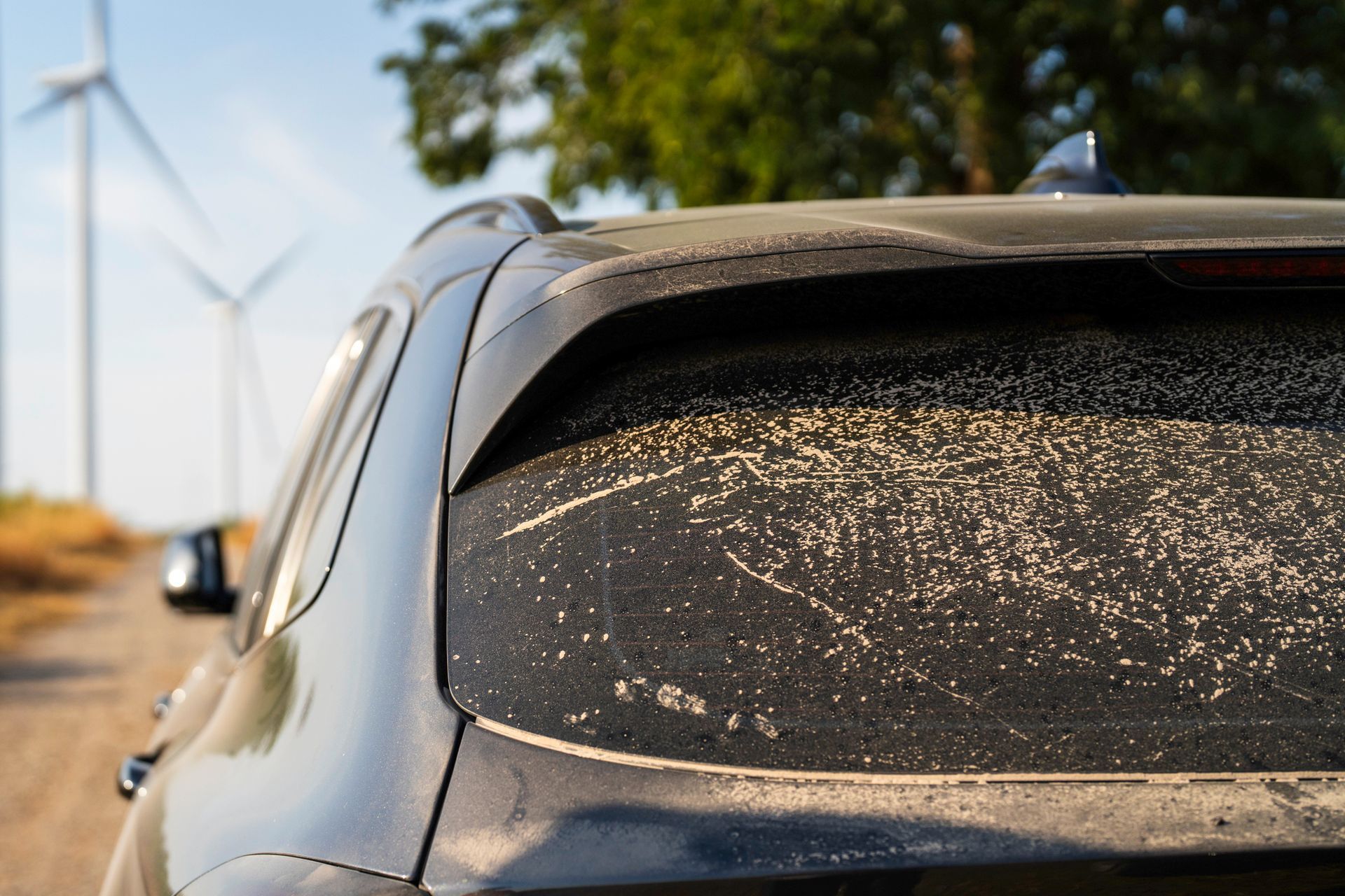 A dirty car is parked on a dirt road with windmills in the background.
