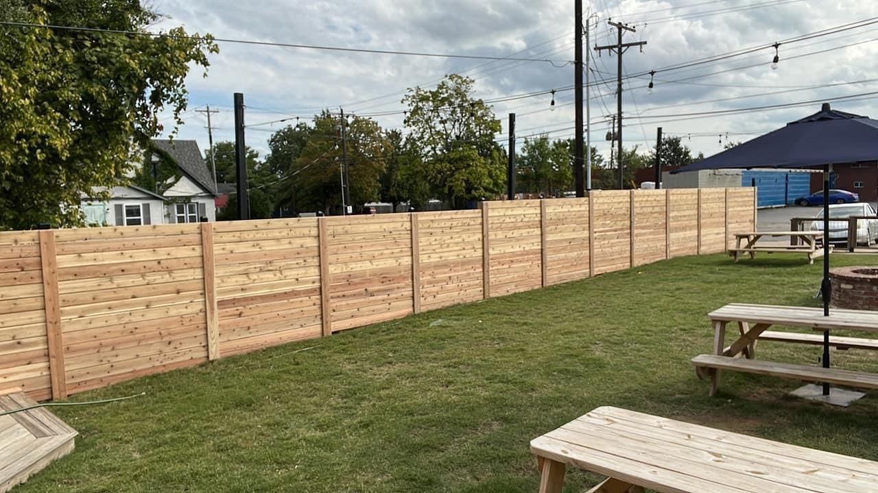 A wooden fence surrounds a grassy yard with picnic tables and umbrellas.