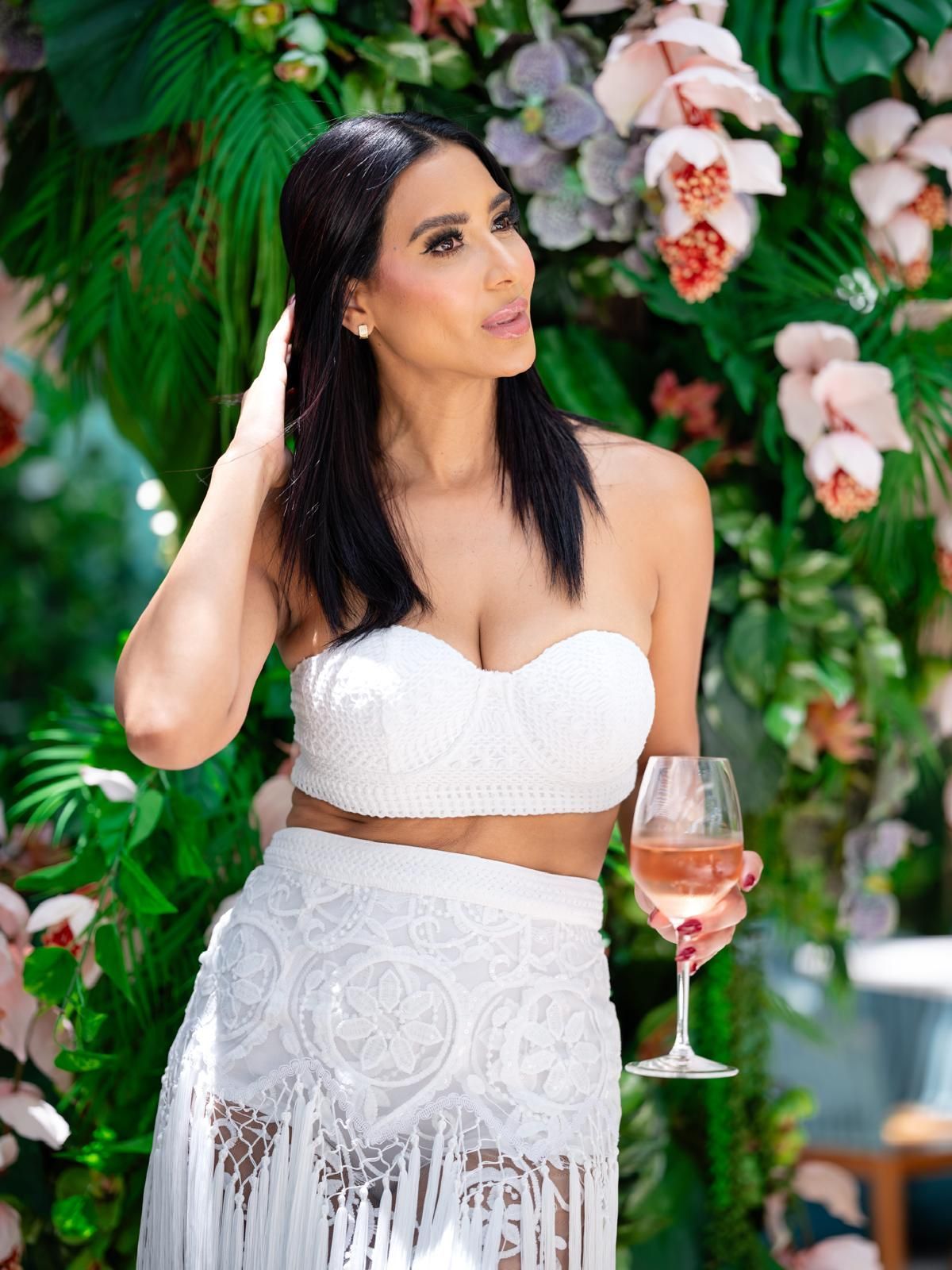 Woman in white outfit holds a wine glass, surrounded by flowers.
