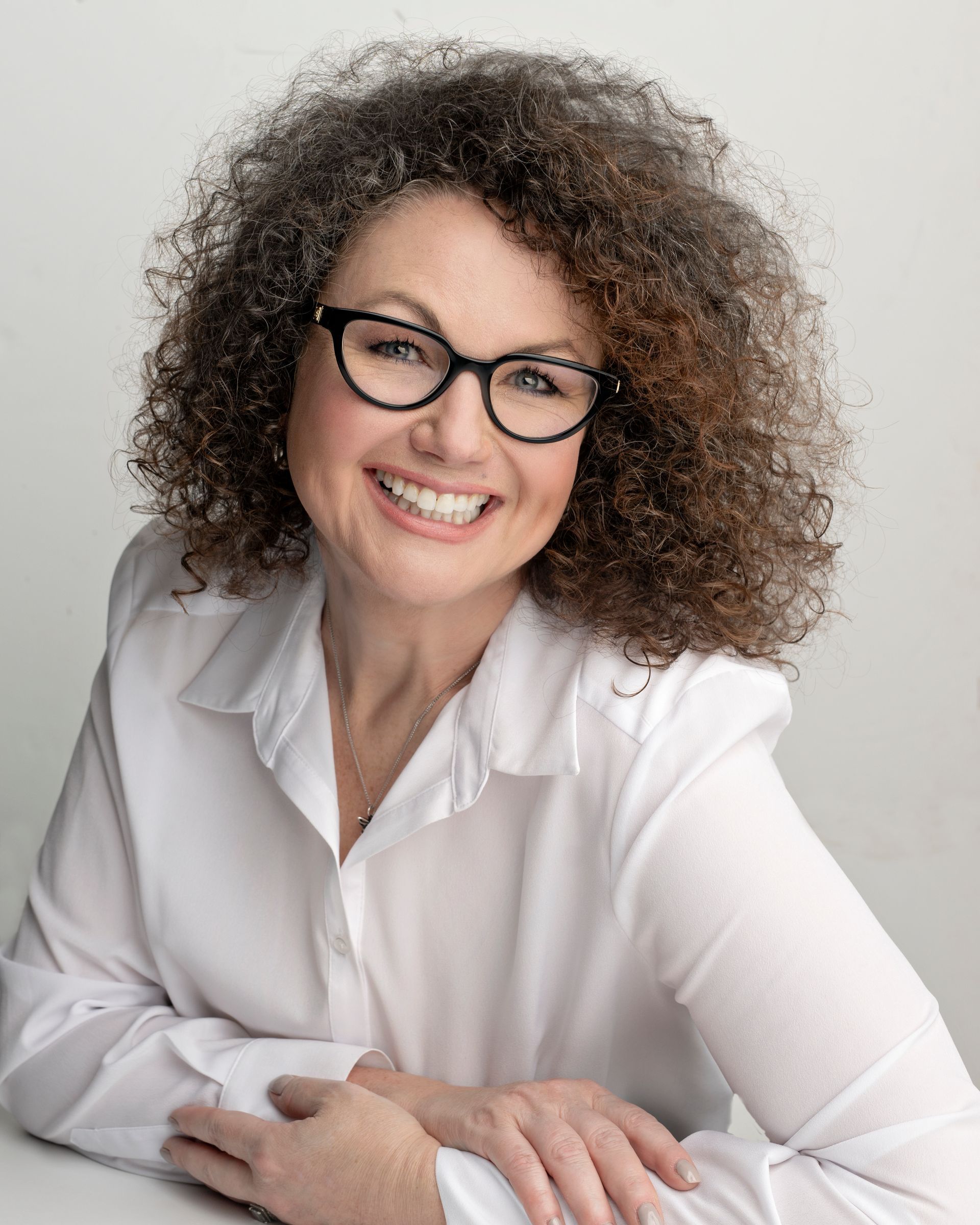 A woman with curly hair wearing glasses and a white shirt is smiling.