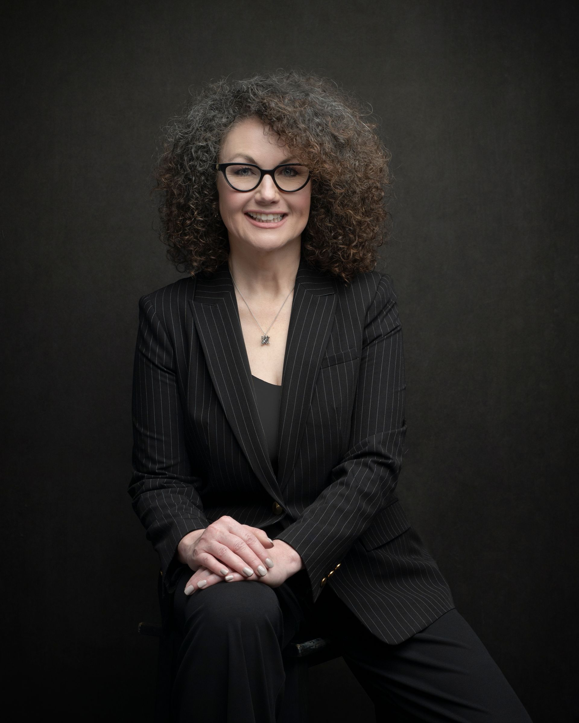 A woman in a black suit and glasses is sitting on a chair.