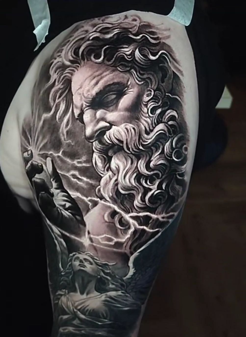 Tattoo of Zeus with lightning and a lower figure. Black and gray shading with detailed curls and a stern expression.