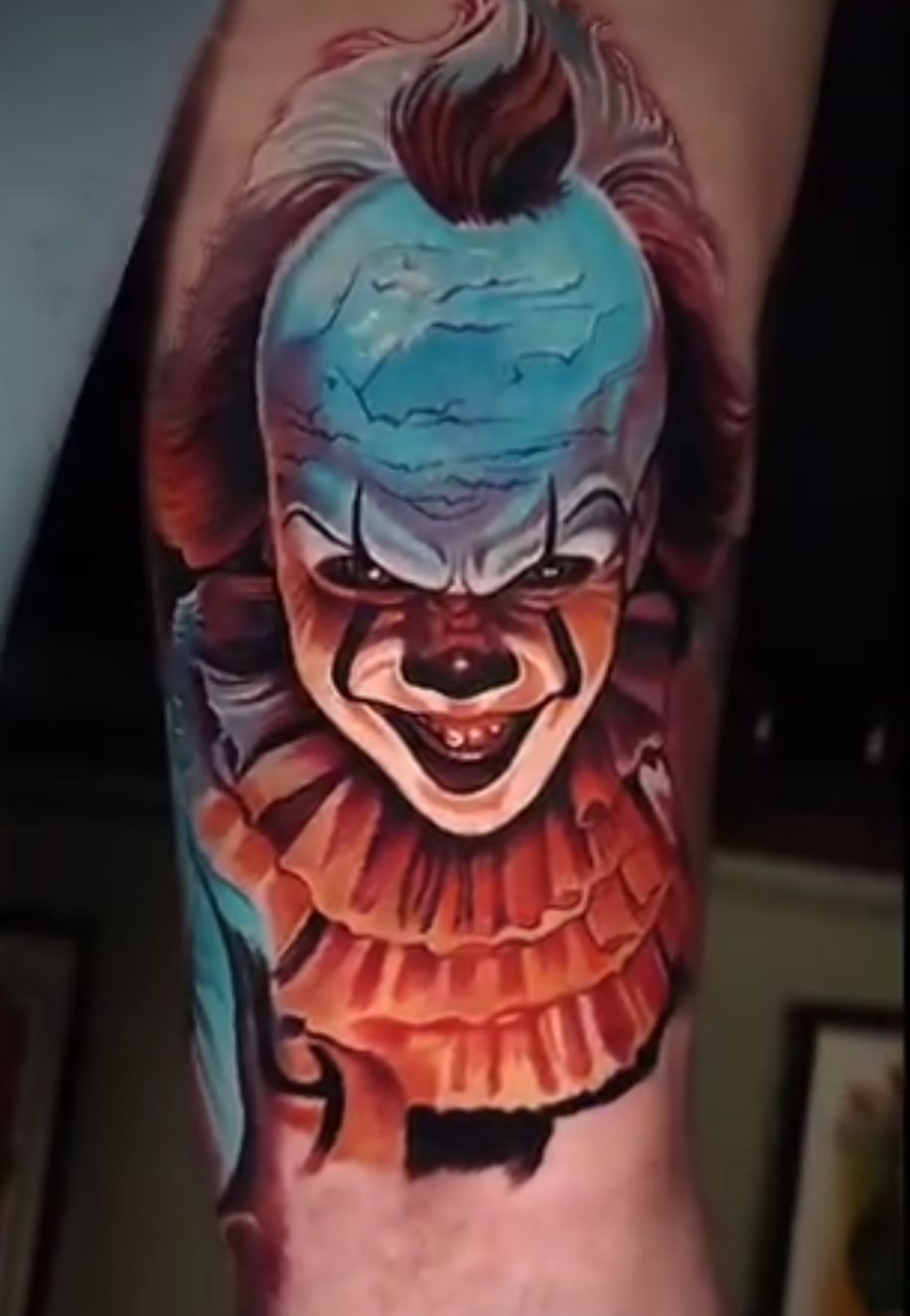 Pennywise the clown tattoo, vibrant colors, evil grin, red ruffled collar, blue and red hair, on a leg.