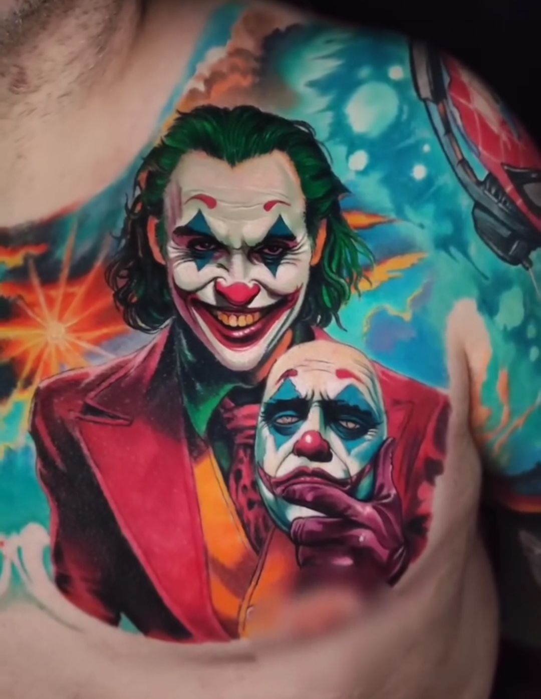 A Joker chest tattoo featuring two portraits. One smiling, holding a clown mask, with colorful background.