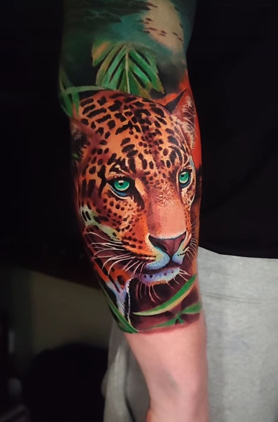 Detailed arm tattoo: Leopard portrait with teal eyes, surrounded by greenery.