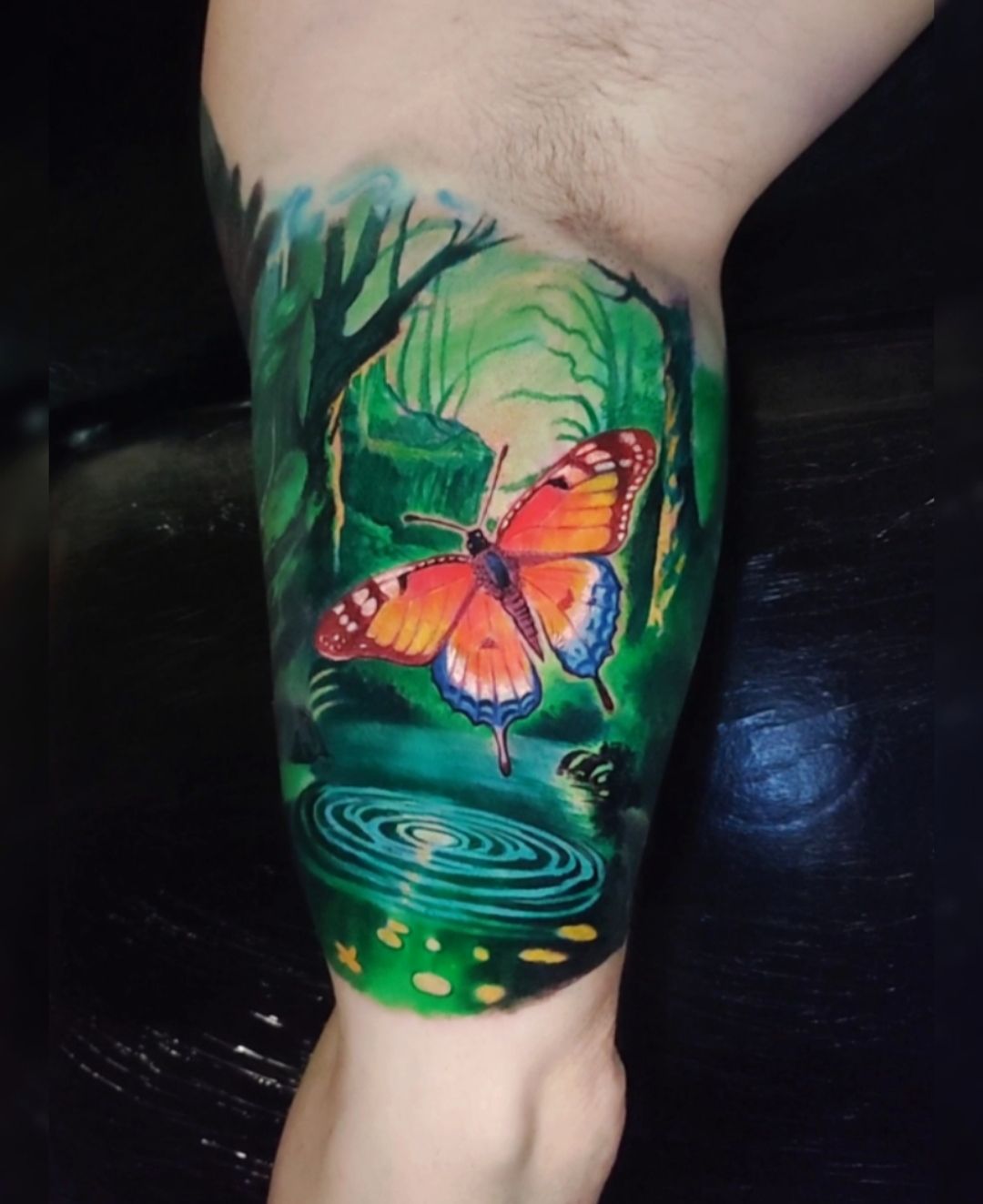 Colorful butterfly tattoo on a leg. The design includes a forest scene with a pond.